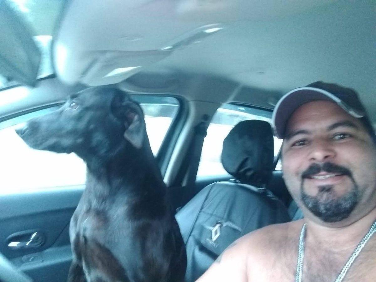 Maurício sorrindo no banco do motorista com Anita, cachorra preta, sentada no banco do carona olhando pela janela.