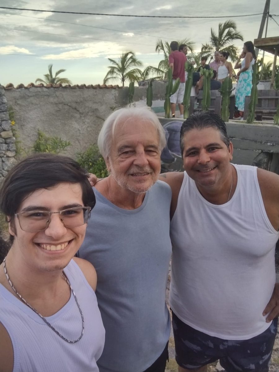 Daniel, o vovô Carlinhos e Maurício abraçados ao fim da tarde, com coqueiros ao fundo.