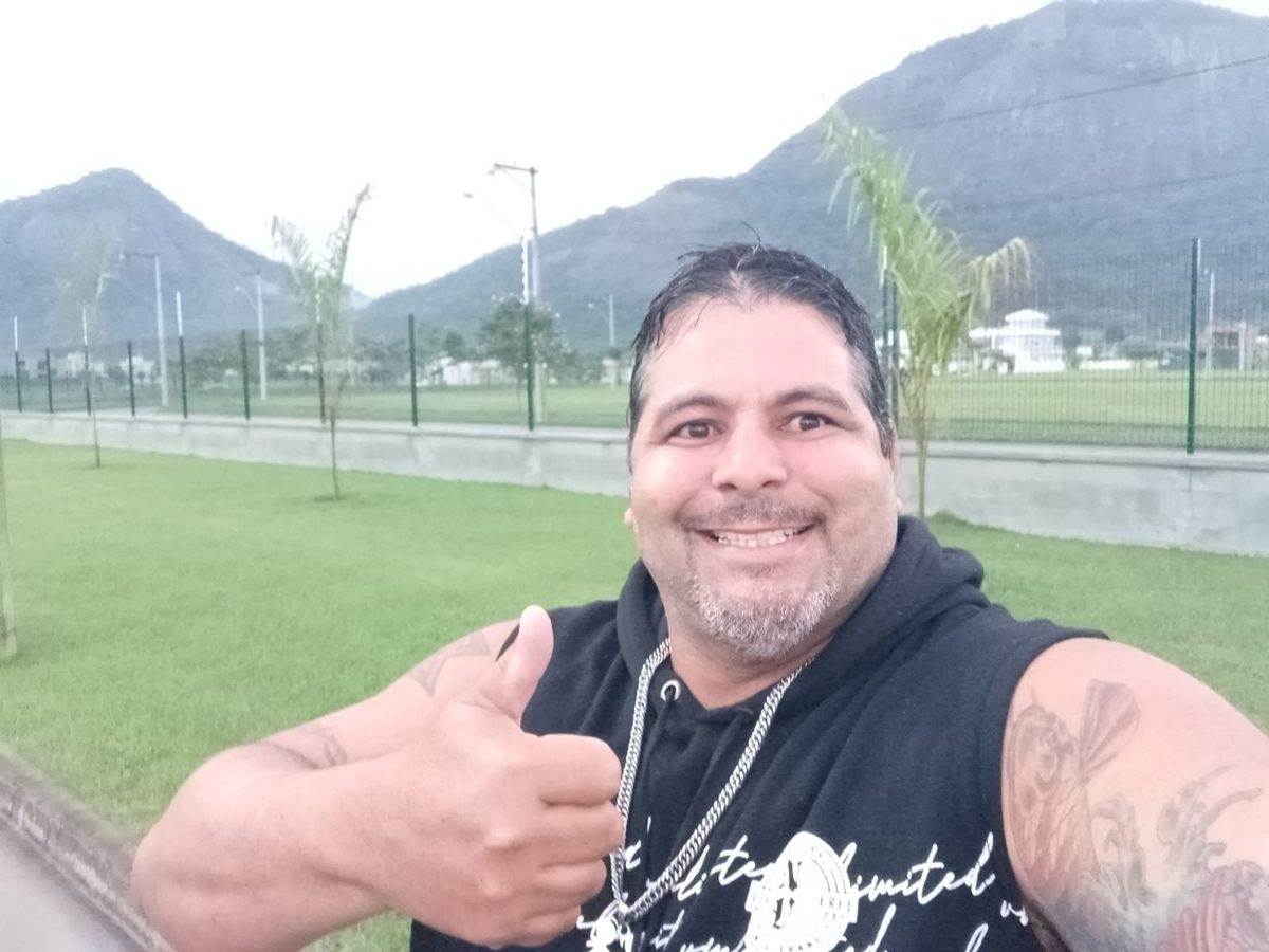 Maurício de regata, polegar para cima, com a serra de Maricá ao fundo.