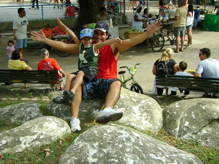 Maurício sentado numa pedra no parque com Daniel pequeno no colo, os dois de braços abertos.