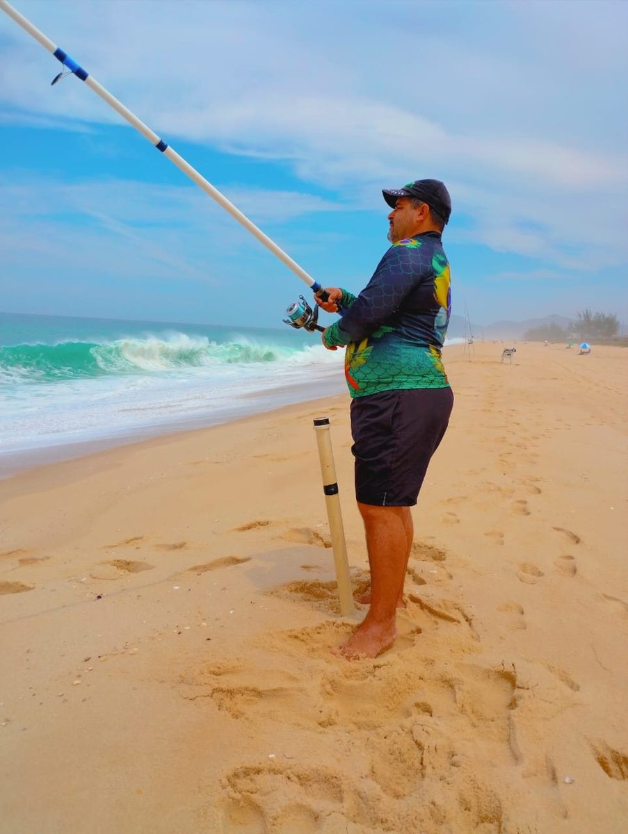 Maurício de pé na areia, vara de pescar erguida, olhando a arrebentação.