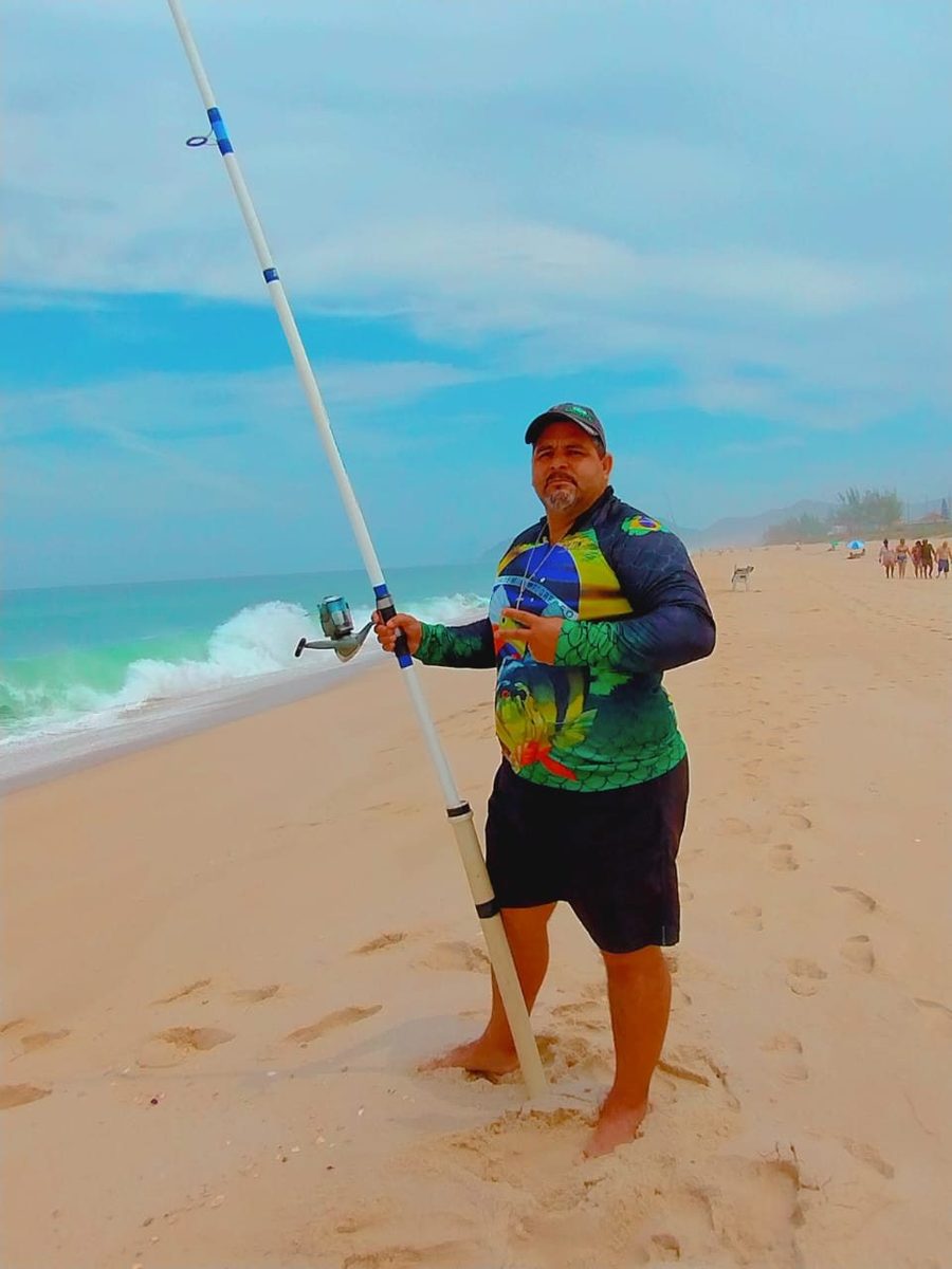 Maurício pescando na beira do mar, camisa de pesca colorida e vara apontada para a arrebentação.