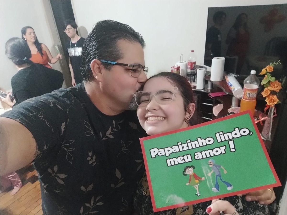 Maurício e Débora sorrindo, segurando uma placa escrita Papaizinho lindo, meu amor.