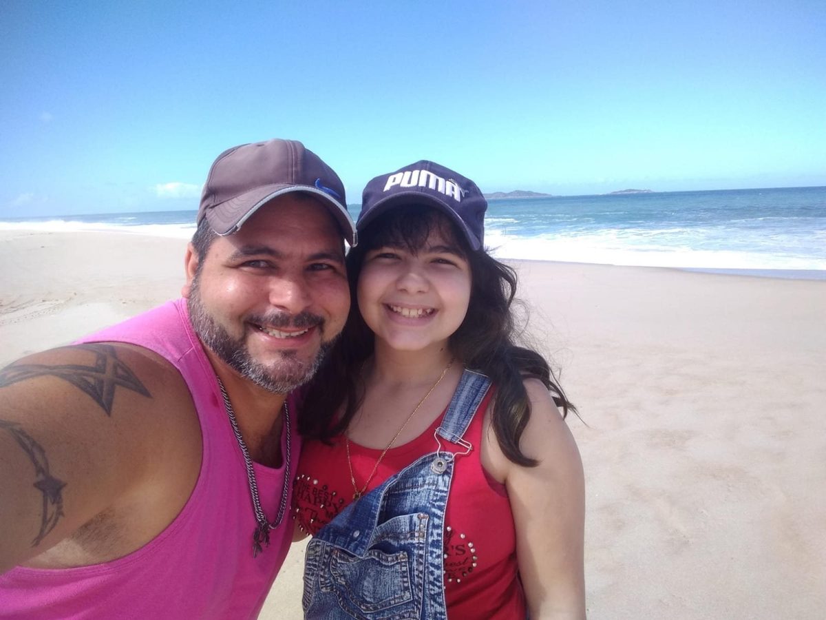 Maurício e Débora pequena na praia, os dois de boné, sorrindo para a câmera.